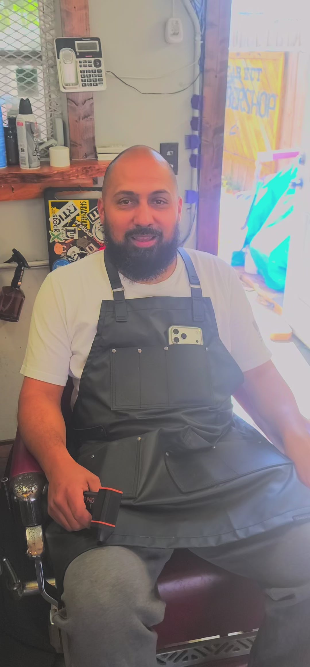 Load video: A barber stands inside a barbershop holding a cordless air duster called Salon Air Pro. He demonstrates how it quickly blows loose hair off a client’s neck and shoulders after a haircut. The barber explains that the tool is lightweight, powerful, and helps keep clients clean without needing a bulky air compressor. The shop environment is active, with barber chairs, mirrors, and tools visible in the background.